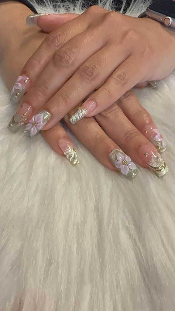 Pink and gold acrylic nails featuring 3D floral designs, glitter accents, and elegant decorative details.